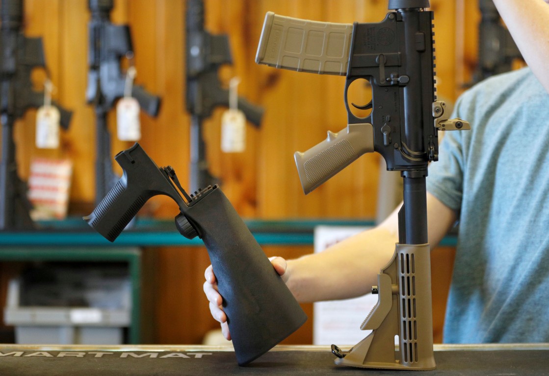 A bump fire stock (L), which attaches to a semi-automatic rifle to increase the firing rate, is...