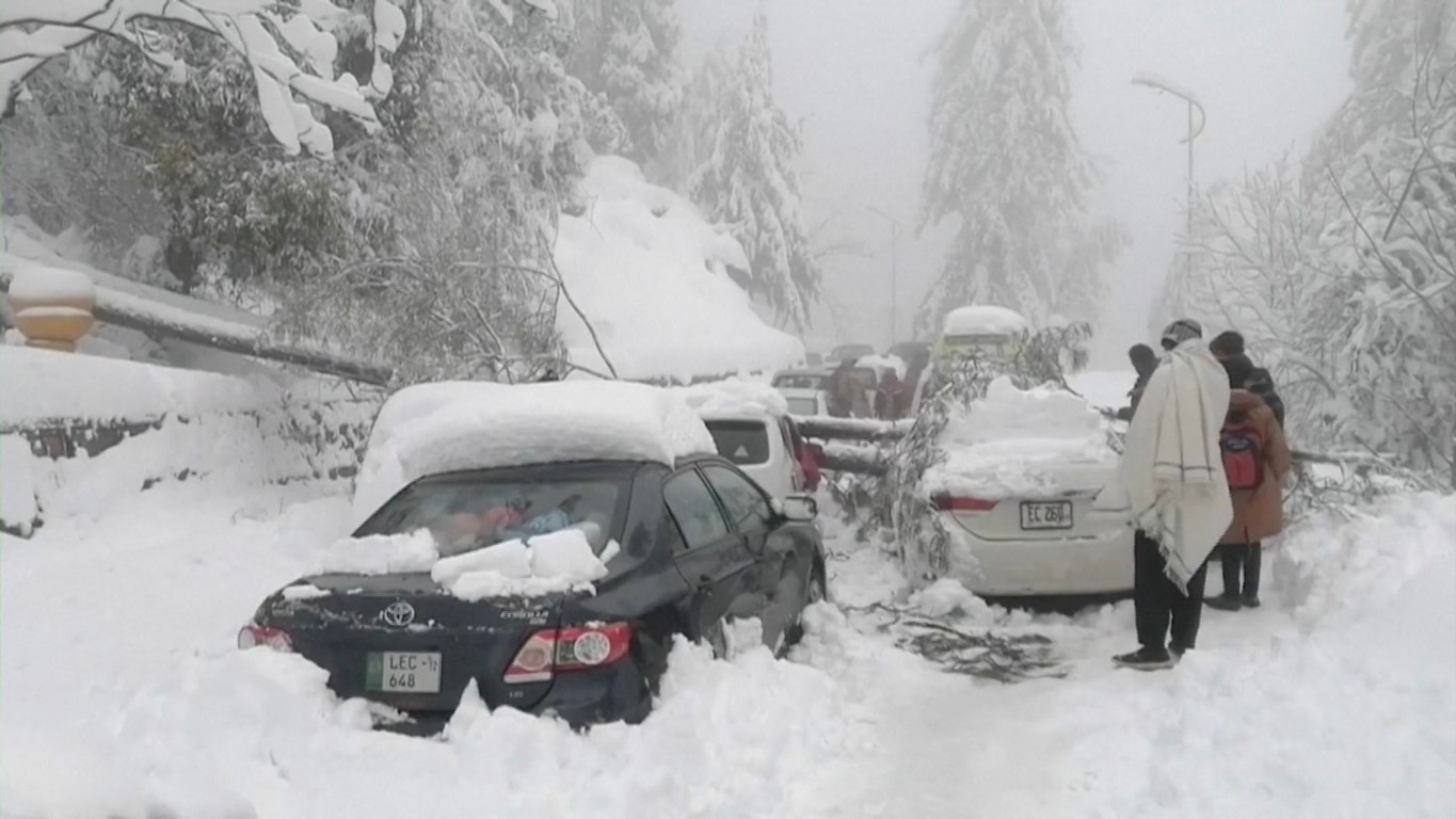 People stand next to cars stuck under fallen trees on a snow-covered road in Murree, northeast of...