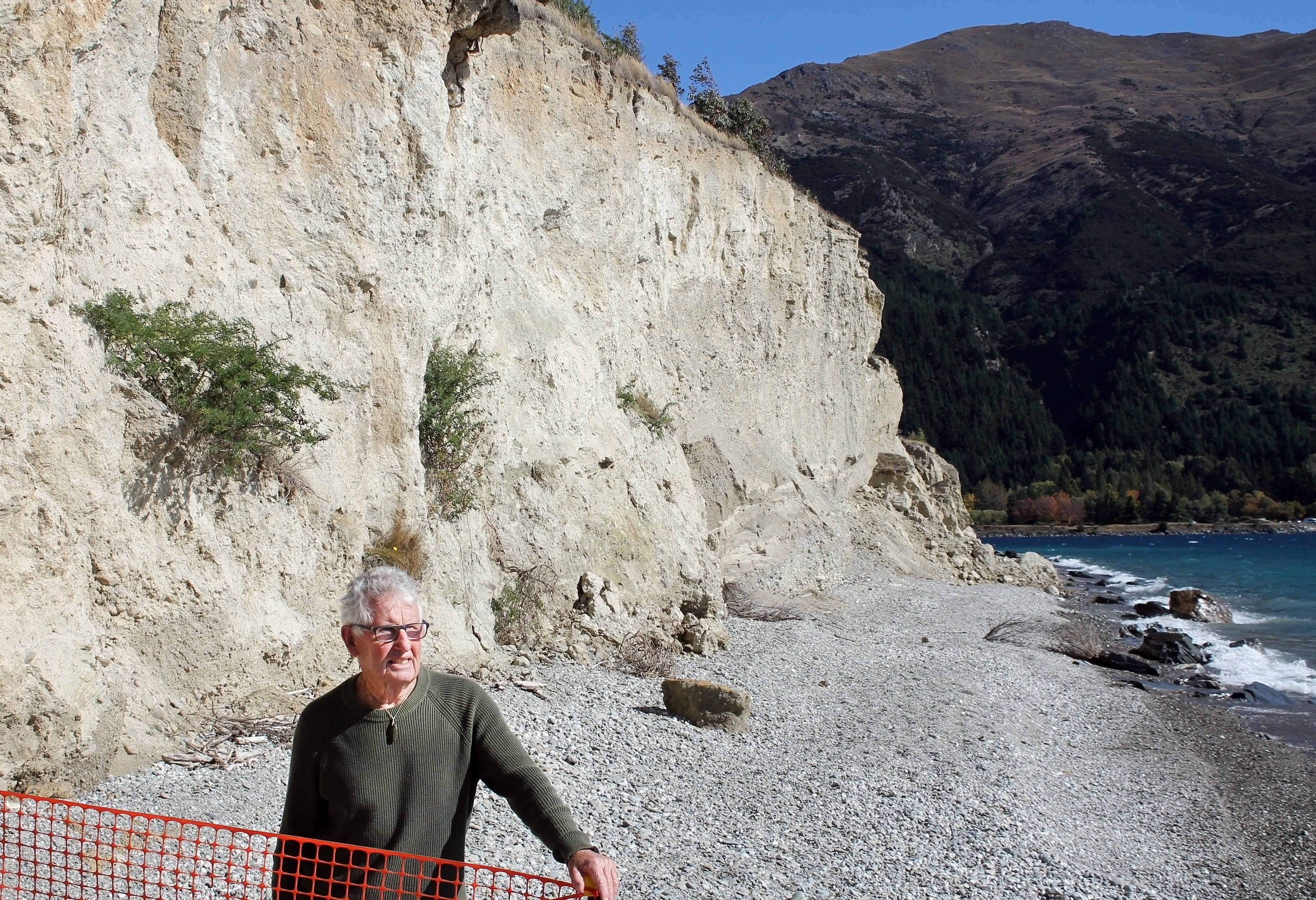 Dave Currie stands in front of the Flora Dora Cliffs at  Lake Hāwea, resting his hand on a flimsy...
