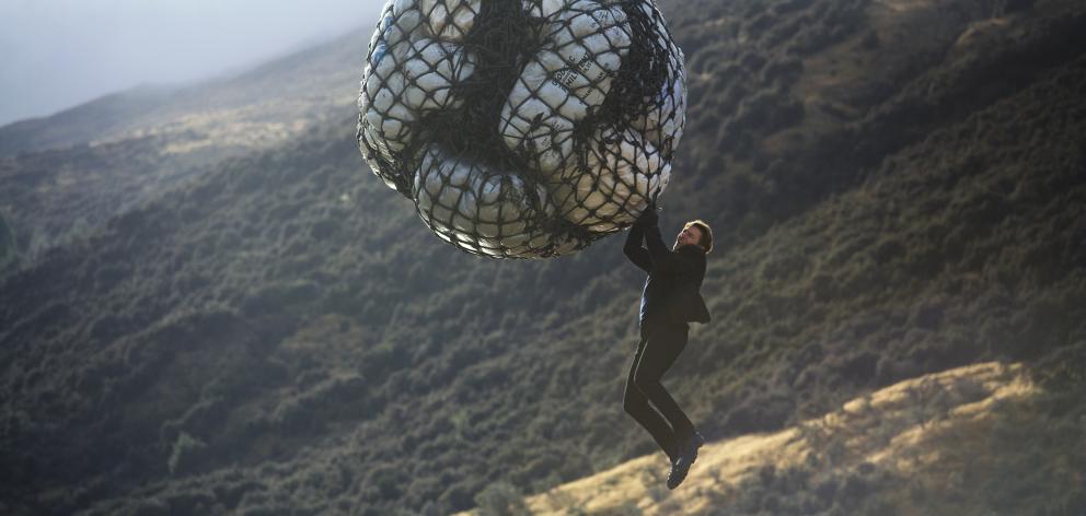 Actor Tom Cruise, as Ethan Hunt in Mission:  Impossible — Fallout, dangles from a bag, attached...