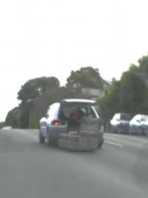 A video still of a man sitting on a couch clinging to the back of a hatchback towing him around...