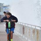 Two boys rider a Lime e-scooter at St Clair as waves crash against the Esplanade wall. Photo: Peter McIntosh