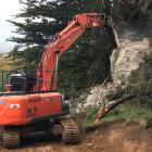 An excavator equipped with a rock-breaking attachment breaks up a large unstable boulder that...