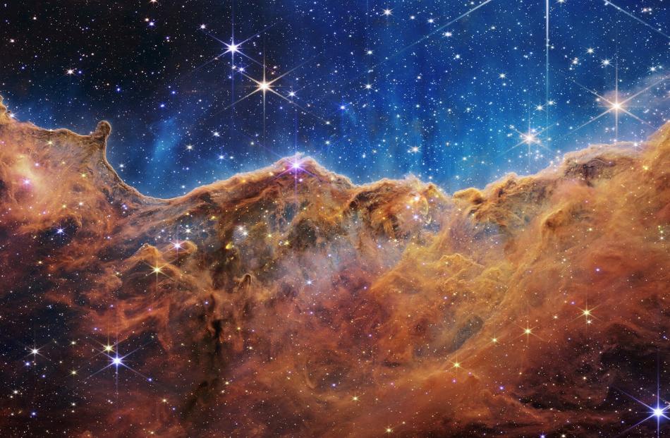 The Cosmic Cliffs of the Carina Nebula are seen in an image divided horizontally by an undulating...